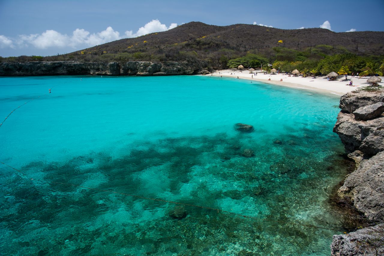services-03 Stunning view of a tropical beach with turquoise waters, rocky shoreline, and sunbathers along the sandy shore.